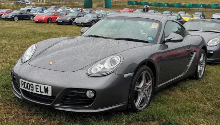 Grey 987 Porsche Cayman coupe parked on grass, side and front three-quarter view