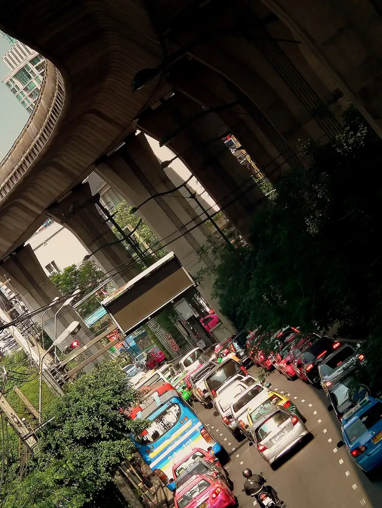 Cars, taxis, and a bus in traffic on Sukhumvit Road in Bangkok, beneath the elevated Skytrain tracks