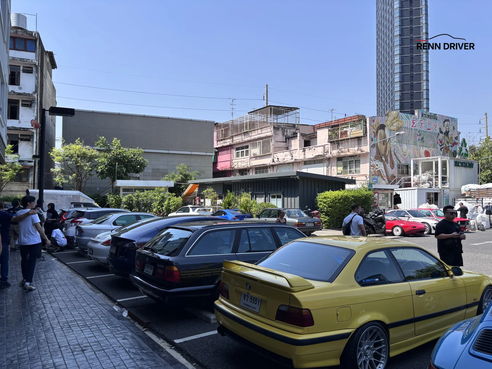Rows of cars parked at a morning meet with a container building and city tower behind